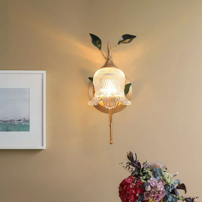 Gold flower wall sconce with clear glass shade on a beige wall, near a framed picture and a bouquet of flowers.