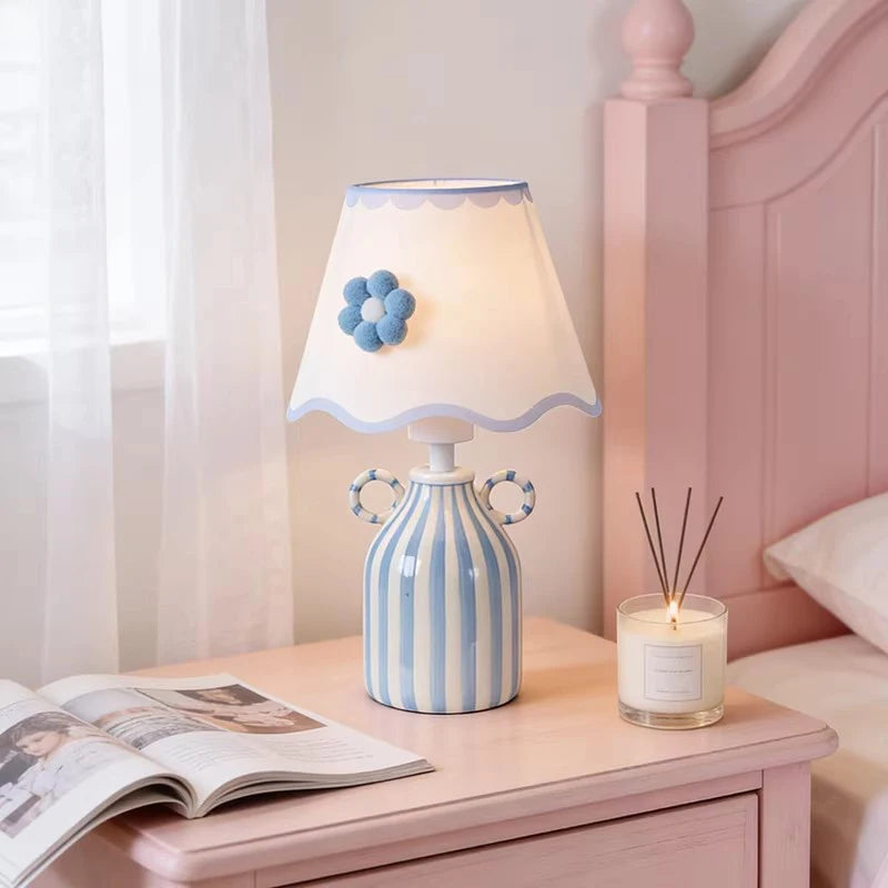 Blue and white striped ceramic table lamp with a scalloped white fabric shade featuring a blue flower accent, placed on a pink nightstand next to a bed with a candle.