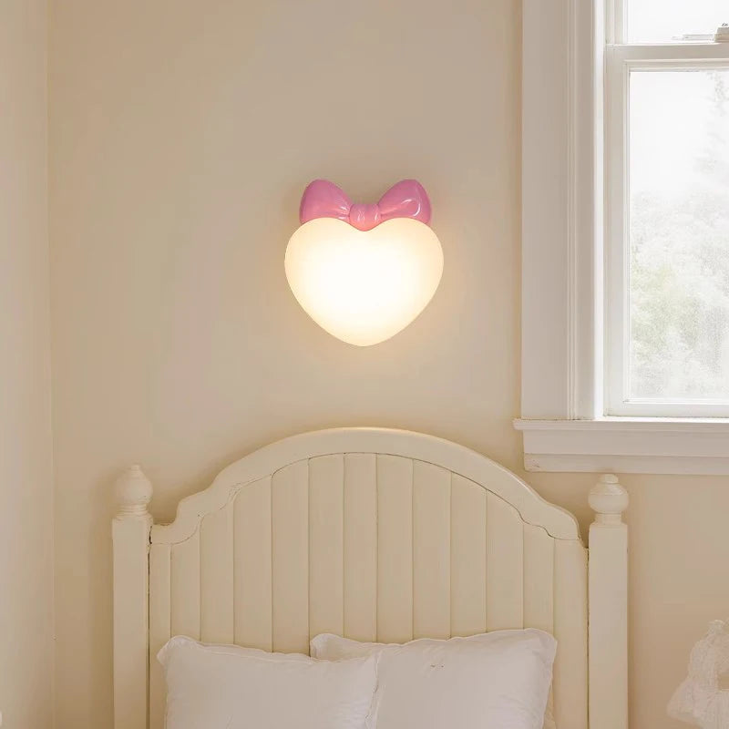 A pink bow heart-shaped wall sconce mounted on a cream-colored wall above a white headboard.