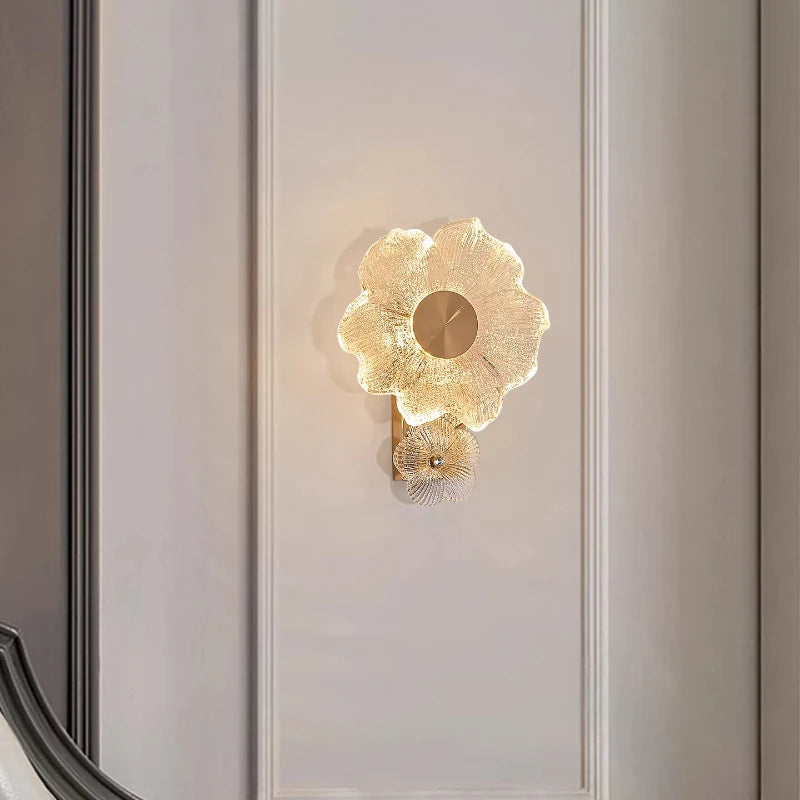 A gold flower-shaped wall sconce with textured glass petals and a central metallic disc, mounted on a white paneled wall.