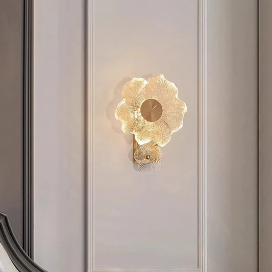 A gold flower-shaped wall sconce with textured glass petals and a central metallic disc, mounted on a white paneled wall.