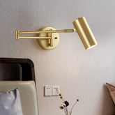 A gold adjustable arm wall sconce mounted on a textured gray wall above a bed, with a light on.