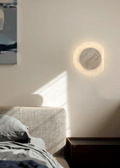 Round wall light fixture on a white wall above a bed with soft lighting.