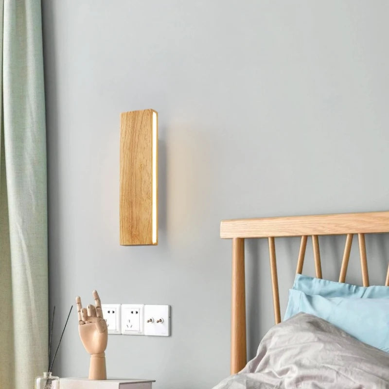 Wooden wall sconce mounted on a light grey wall next to a bed with blue pillows and a wooden headboard, showing electrical outlets below.