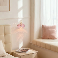 A single pink faceted crystal pendant light with a gold accent hanging above a nightstand in a bedroom setting.