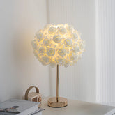 A white floral rose bud table lamp with a gold base and stem, illuminated on a white surface.