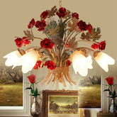A close-up view of a gold floral pendant light with red rose buds, green leaves, and frosted white tulip-shaped shades against a beige wall.
