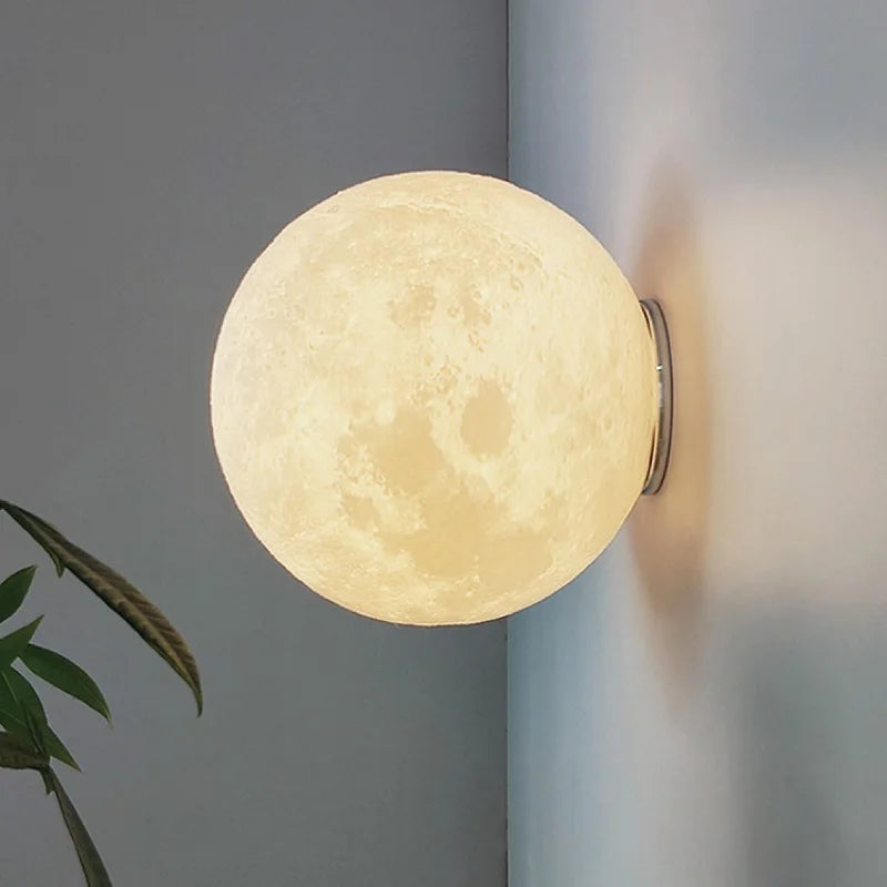 A 3D printed moon shaped wall sconce with a textured surface is mounted on a gray wall, emitting a soft yellow light. A green leaf plant is visible in the foreground.