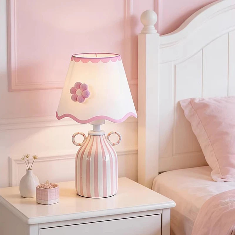 Pink and white striped ceramic table lamp with a scalloped white fabric shade featuring a pink flower accent, placed on a white nightstand next to a bed.