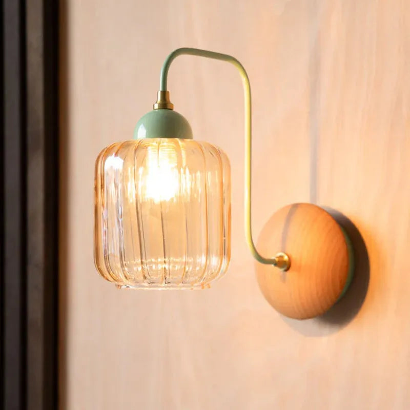 A wall sconce with a green arm, a clear ribbed amber glass shade, and a round wooden backplate, mounted on a light-colored wood-paneled wall.