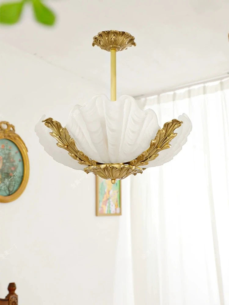 White frosted seashell glass pendant light with ornate gold leaf accents and a central brass stem, hanging against a soft white curtain with framed art on the wall.