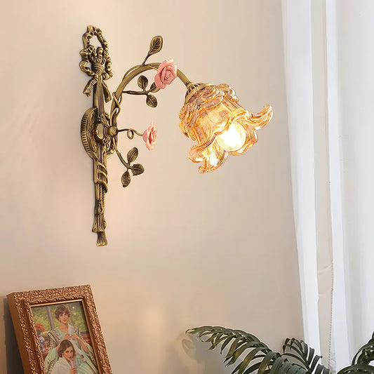 Antique gold flower wall sconce with a peach-tinted glass shade and pink rose accents, mounted on a light-colored wall with a framed picture below.