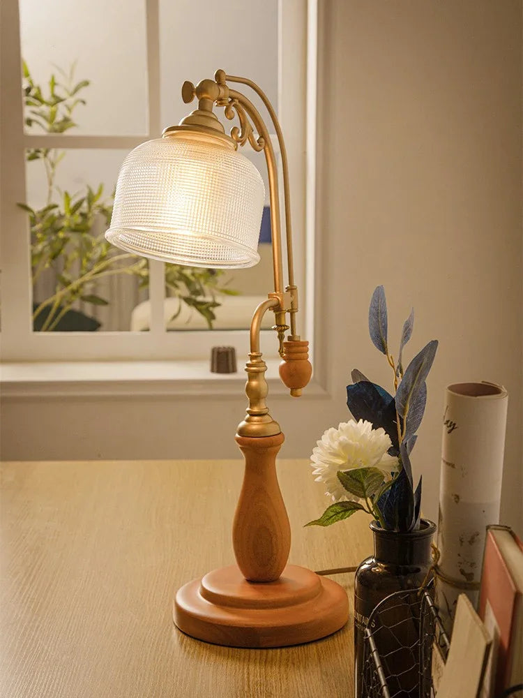 Close-up of a wooden base table lamp with a textured glass shade, featuring an antique brass finish arm and a knob switch, with a vase of flowers in the background.