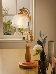 Close-up of a wooden base table lamp with a textured glass shade, featuring an antique brass finish arm and a knob switch, with a vase of flowers in the background.