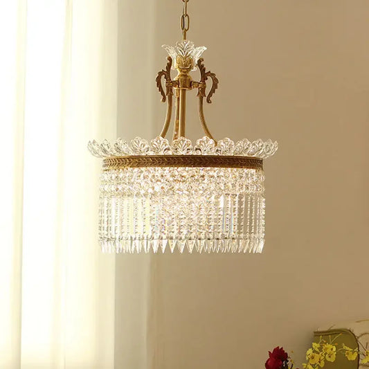 A gold and crystal tiered chandelier with cascading teardrop crystals, shown hanging in a room with sheer curtains.