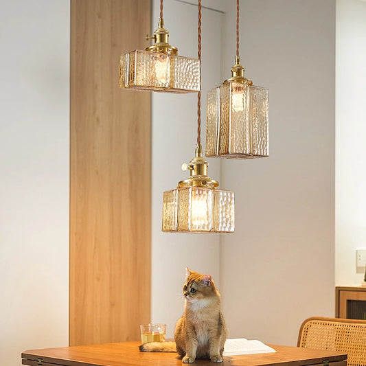 Three glass square pendant lights with brass fixtures hanging above a wooden table with a cat.