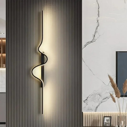 A modern gold wall sconce with a wavy design mounted on a ribbed gray wall, emitting a warm glow.