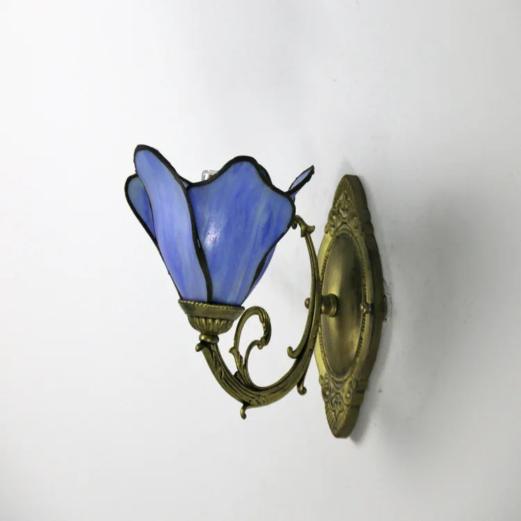 A blue Tiffany-style wall sconce with a flower-shaped stained glass shade, attached to a decorative distressed gold metal arm and backplate against a white wall.