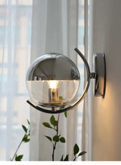 Wall-mounted lamp with glass shade and metal arm in a room with curtains and plants.