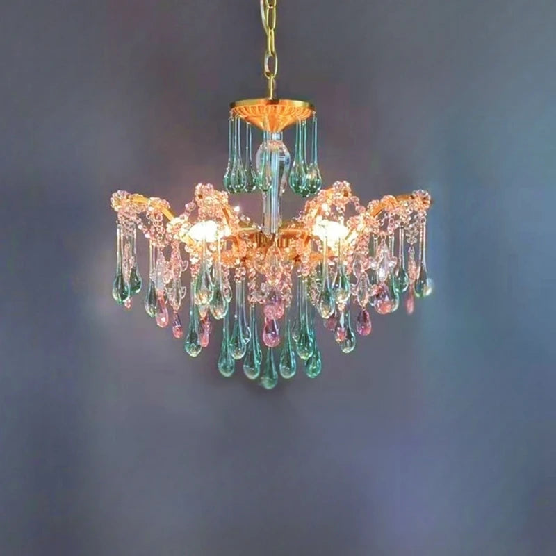Close-up of a gold chandelier featuring numerous teardrop crystals in pink and green hues against a dark blue wall.