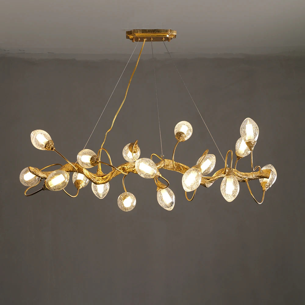 A linear gold chandelier designed to resemble a tree branch with clear, textured glass globes, suspended against a plain dark background.