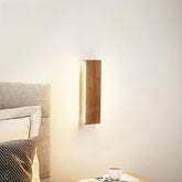 Wooden wall sconce mounted on a white wall next to a bed with pillows and a bedside table with a cup of coffee.