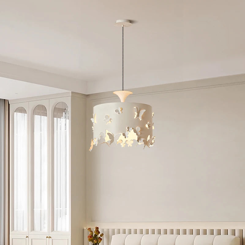 A cream-colored drum pendant light with butterfly cutouts, shown in a room with white curtains and furniture. The light illuminates the surrounding space.