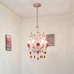A pink and gold chandelier with rose and crystal details hangs from the ceiling in a room with white walls and decorative artwork.