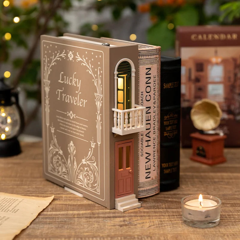 A brown bookend lamp titled 'Lucky Traveler' with a balcony and illuminated window, placed on a wooden surface next to other books and a candle.
