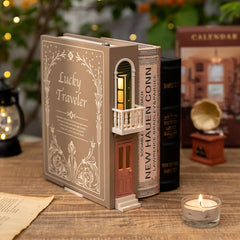 A brown bookend lamp titled 'Lucky Traveler' with a balcony and illuminated window, placed on a wooden surface next to other books and a candle.