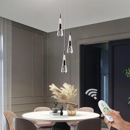 Three gold crystal teardrop pendant lights hanging over a white dining table, a remote control is visible in the foreground.