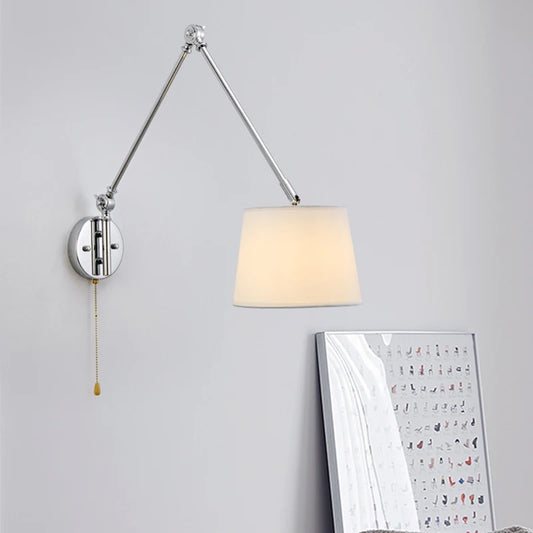 Chrome swing arm wall sconce with a white fabric drum shade and a pull chain, mounted on a white wall.