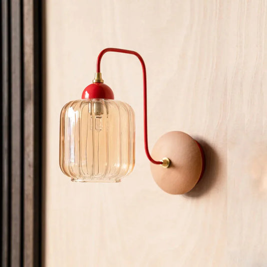 A wall sconce with a curved red arm, a clear ribbed amber glass shade, and a round wooden backplate, mounted on a light-colored wood-paneled wall.