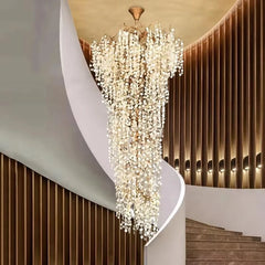 A large, cascading chandelier with numerous clear crystal teardrops and a gold-toned metal frame hangs in a grand entryway with a curved staircase.