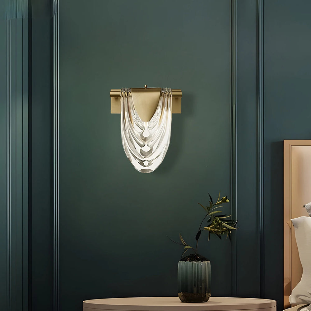 A gold wall sconce with a clear glass shade, installed on a dark teal wall above a side table with a potted plant.