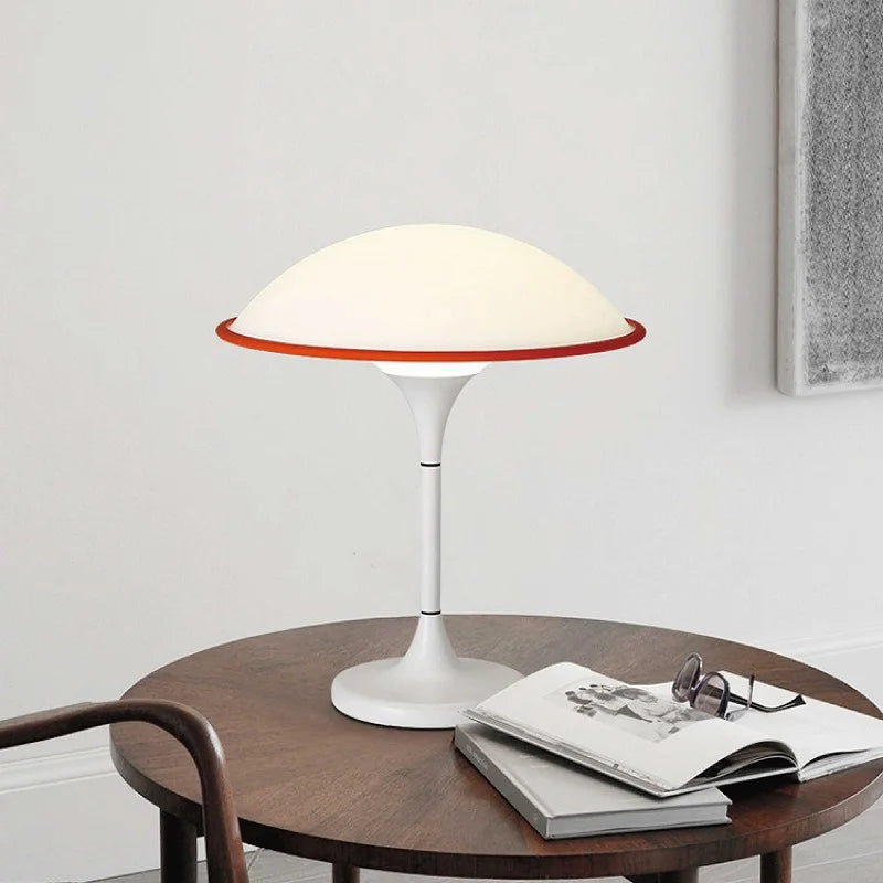 White dome table lamp with a red accent ring, placed on a round wooden table next to an open book and eyeglasses.