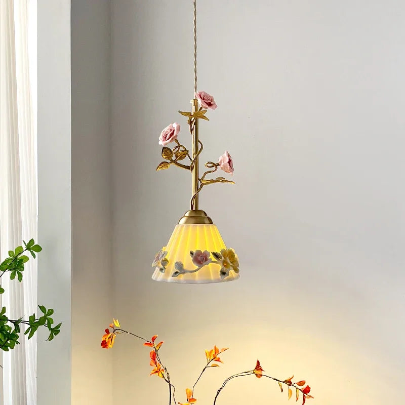 A single pendant light with a brass stem, pink rose buds, and a frosted glass shade with floral patterns, hanging against a white wall.