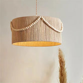 Close-up of a cylindrical pendant light fixture wrapped in natural-colored hemp rope, with a strand of small wooden beads draped along the top edge.