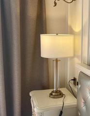Close-up view of a clear glass cylinder table lamp with a brushed gold base and accents, showing the fabric shade and switch.