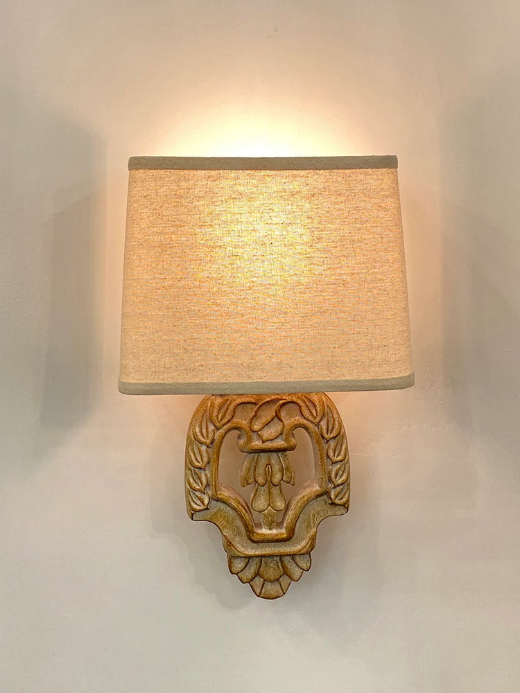 A vintage wood wall sconce with a carved base and fabric shade, illuminated against a white wall.