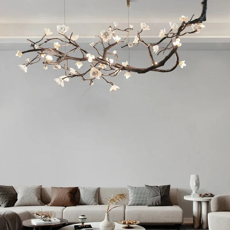 A long, artistic chandelier shaped like a branching tree with white floral accents and illuminated LED lights, hanging over a living room sofa.