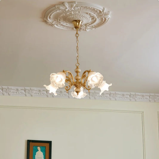A gold chandelier with five frosted glass shades shaped like flowers, hanging from a white ceiling with ornate molding.