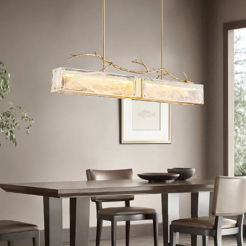 Rectangular glass pendant light with brushed gold branch accents hanging above a dining table.