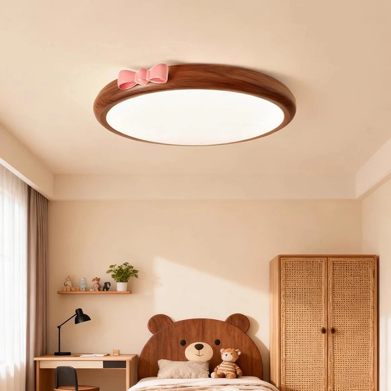 A round, flush mount ceiling light with a wood-tone frame and a pink bow accent, seen in a child's bedroom with a small desk and a wooden cabinet.