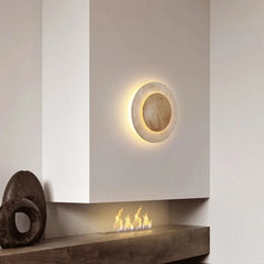 A round wall sconce with a natural stone finish and warm LED light, mounted on a white wall above a modern fireplace with a gas flame.