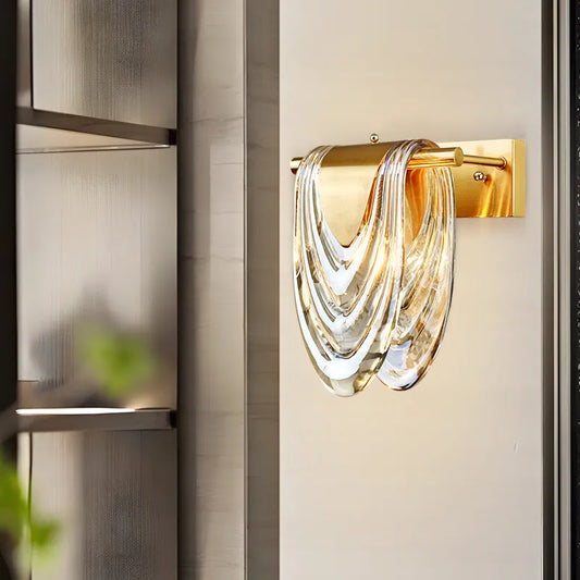 Close-up of a gold wall sconce with a cascading clear glass shade, mounted on a textured wall.