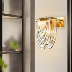 Close-up of a gold wall sconce with a cascading clear glass shade, mounted on a textured wall.