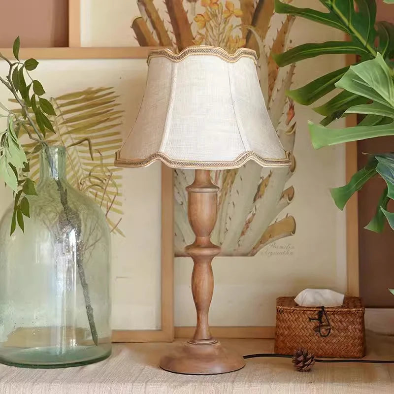 Wooden table lamp with a white shade on a surface with plants and a decorative mirror in the background.