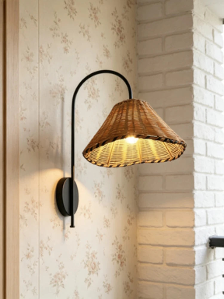 Black metal arm wall sconce with a woven rattan cone shade against a floral wallpaper next to a white brick wall.