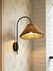 Black metal arm wall sconce with a woven rattan cone shade against a floral wallpaper next to a white brick wall.